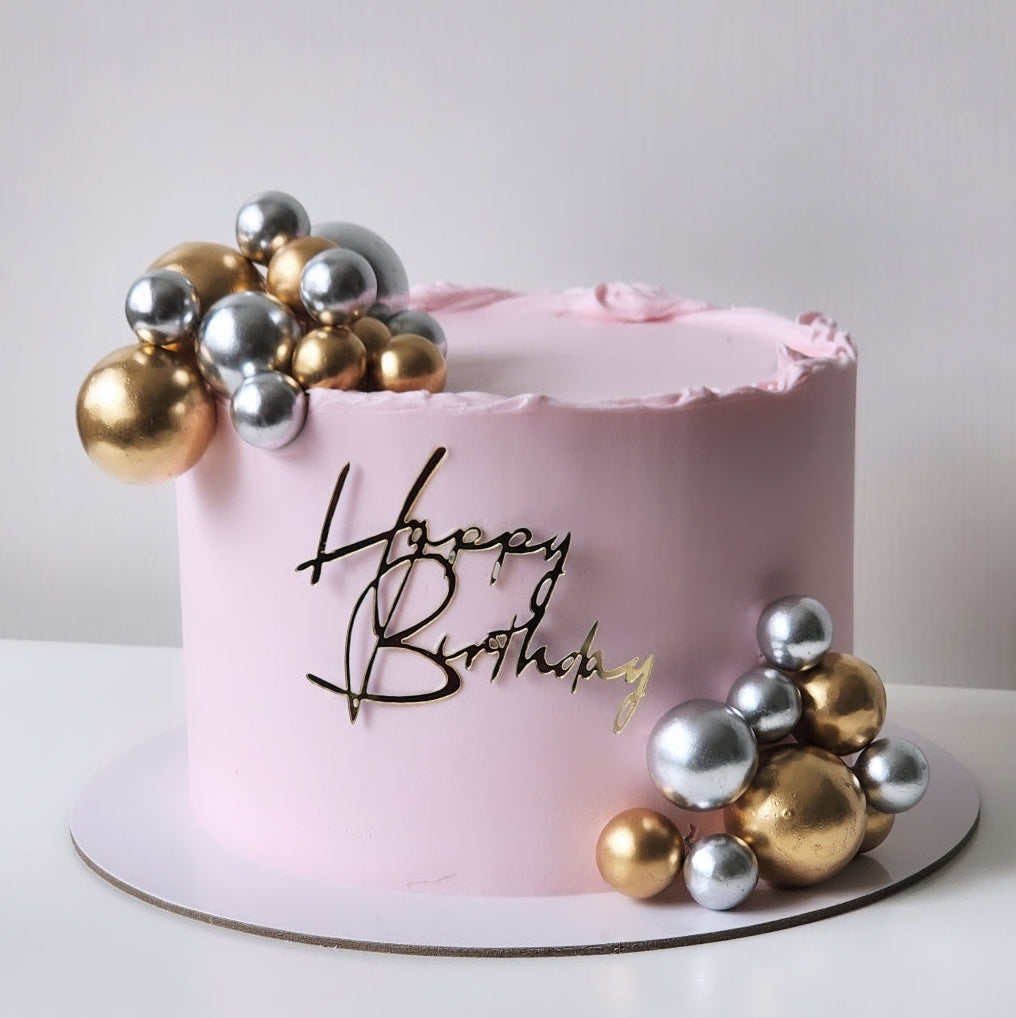 A round pink frosted birthday cake on a white board, decorated with metallic gold and silver balls and a gold 'Happy Birthday' script topper, against a plain light background. Sydney