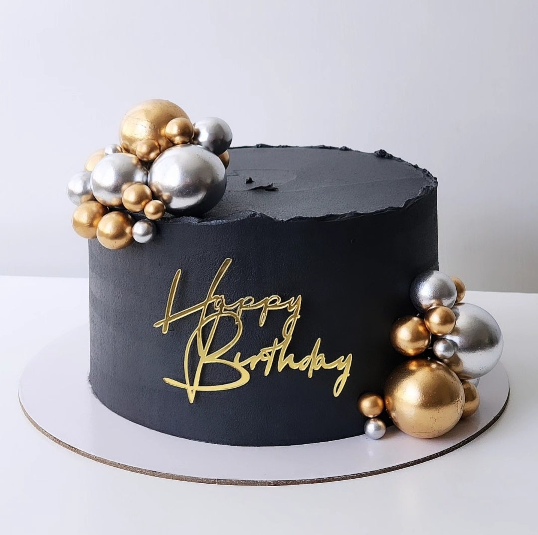 A decorated birthday cake with black icing, golden and silver ornamental balls, and a 'Happy Birthday' message in gold icing.