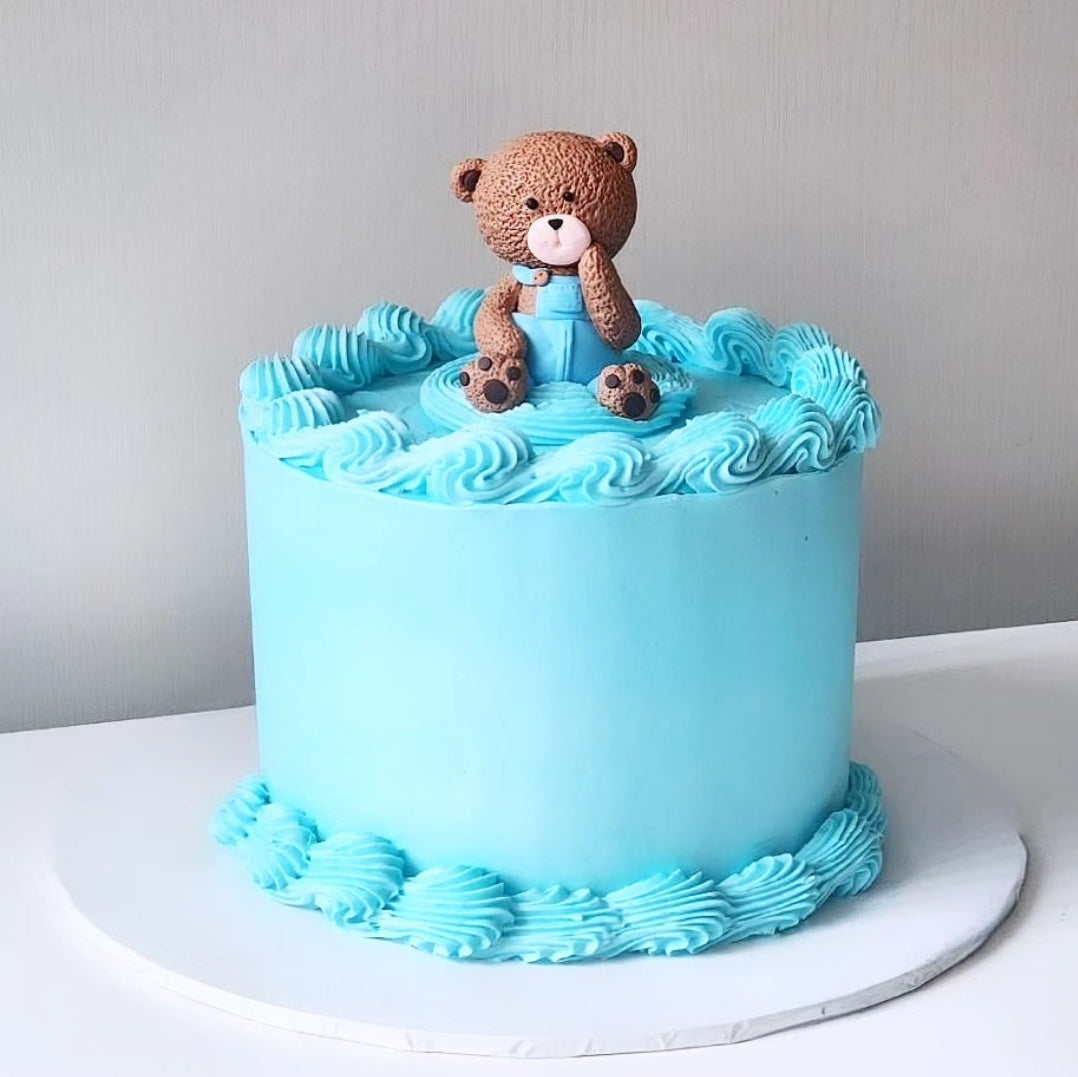 A blue frosted cake with a teddy bear figurine sitting on top, decorated with piped blue buttercream icing.