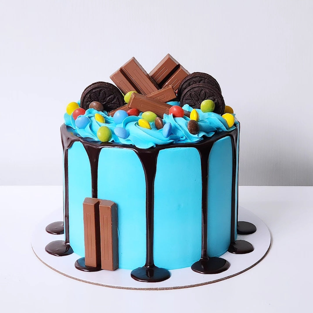 A chocolate cake with a blue buttercream topping and chocolate ganache drip, decorated with an orange and yellow candy piece, on a white cake stand.