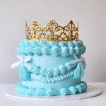 A multi-layered cake with a blue velvety frosting and a golden-colored acylic crown on top, placed on a white cake stand.