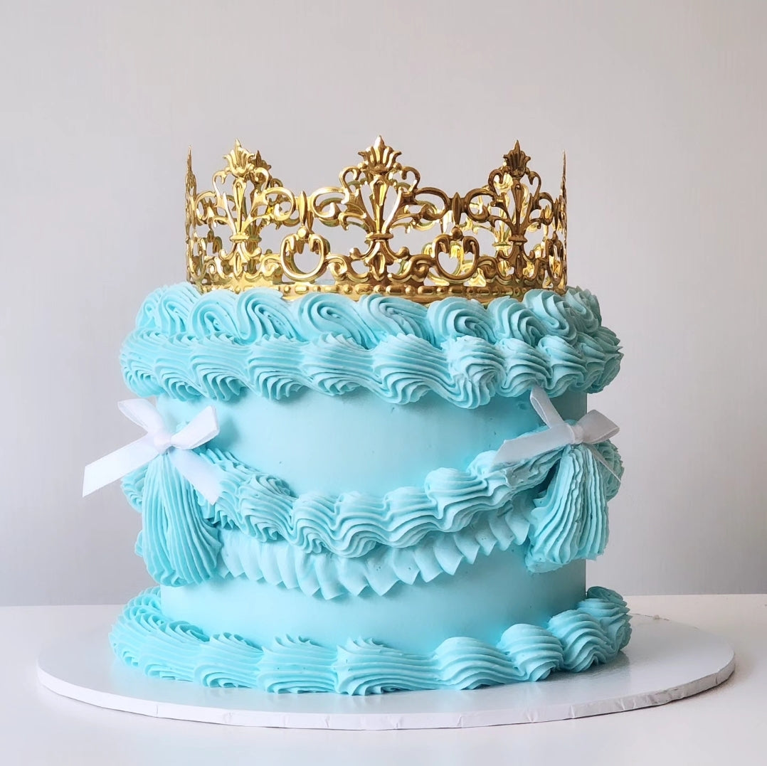 A multi-layered cake with a blue velvety frosting and a golden-colored acylic crown on top, placed on a white cake stand.