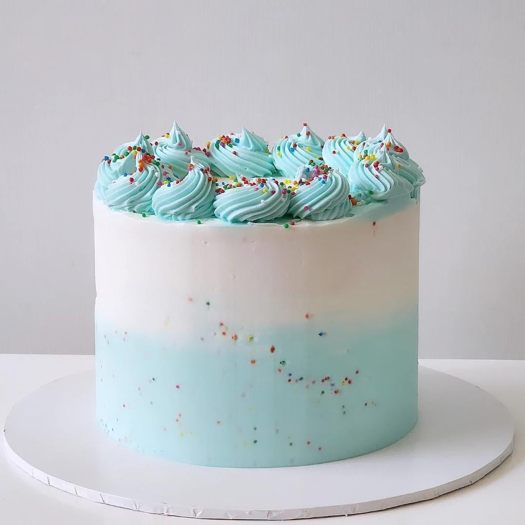A multi-layered cake with a creamy blue frosting and rainbow sprinkles on top, placed on a white round cake stand.