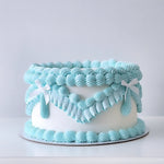 A frosted cake with white and blue icing, featuring a classic vintage design with piped swirls and ribbon detail.