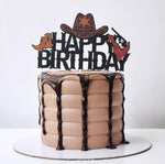 A chocolate cake decorated to look like a cowboy hat and boots with 'HAPPY BIRTHDAY' text, and a cake topper in the shape of a gun.