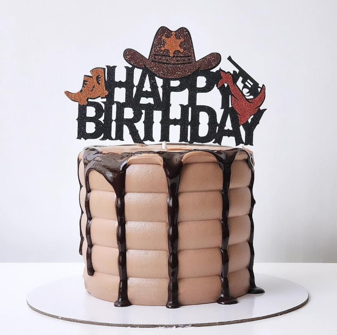 A chocolate cake decorated to look like a cowboy hat and boots with 'HAPPY BIRTHDAY' text, and a cake topper in the shape of a gun.
