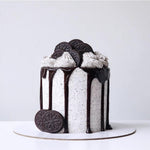 A chocolate cookies and cream cake with a white sponge layers, chocolate glaze, and cookie toppings, set on a white cake stand.