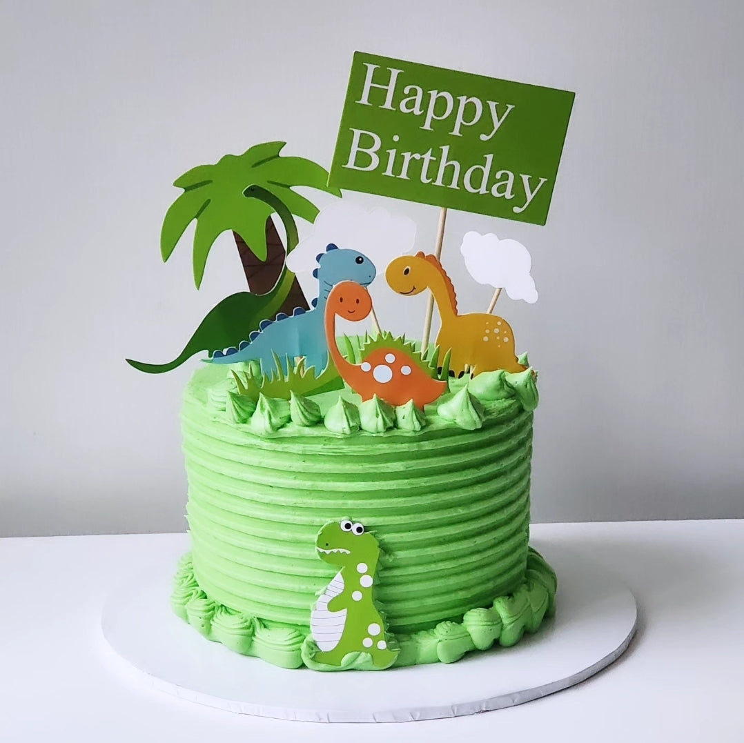 A themed dinosaur cake with a green and white color scheme, featuring cartoonish dinosaur figures and a 'Happy Birthday' sign.