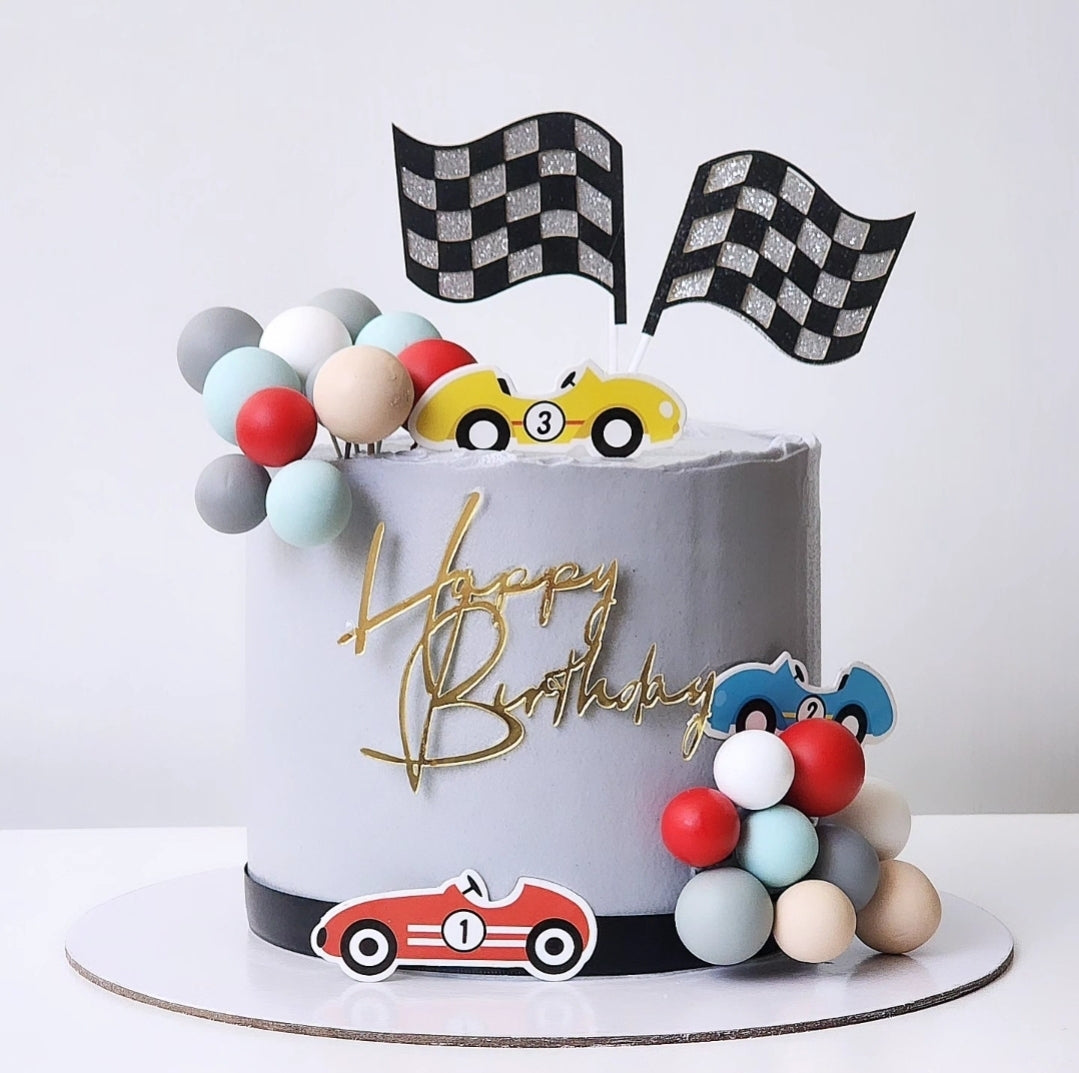 A themed racing car birthday cake with layers decorated to look like cars and checkered flags, frosted with vanilla buttercream and adorned with colorful balls and a 'Happy Birthday' sign.