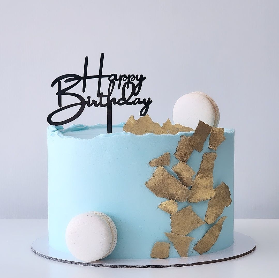 A light blue frosted birthday cake on a white board, decorated with gold leaf flakes, two white macarons, and a black 'Happy Birthday' script topper, sydney 