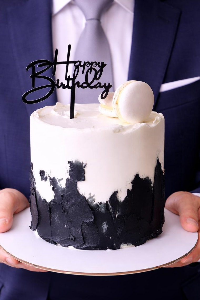 Small black and white birthday cake with a 'Happy Birthday' topper held by a person in a suit.