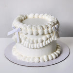 A multi-layered white cake with cream frosting and decorative white piped swirls, topped with a satin ribbon.