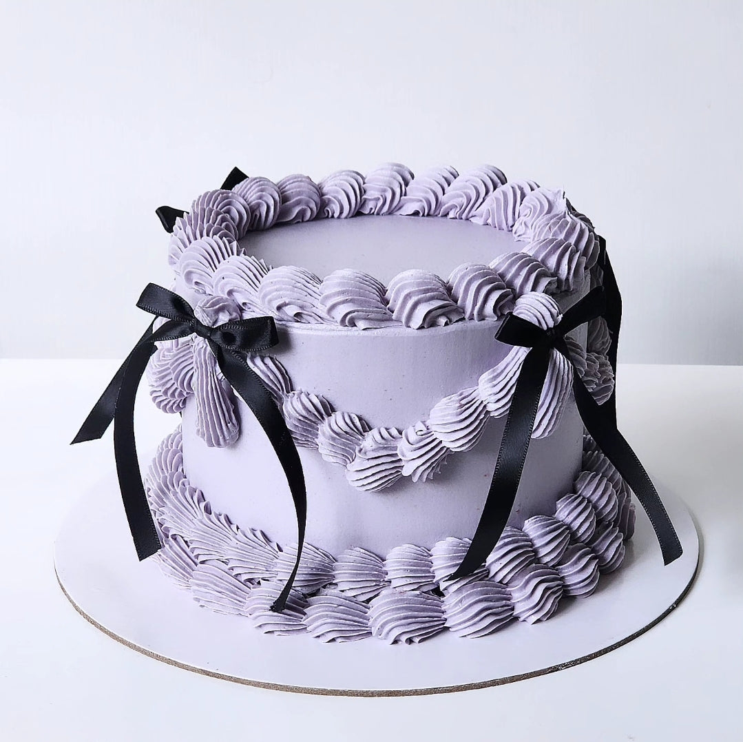 A round cake with a purple buttercream frosting, adorned with a black bow.