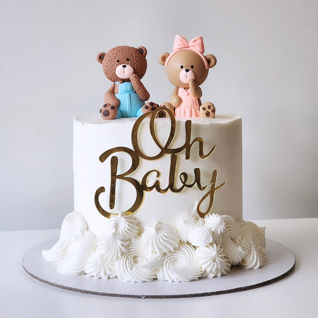 A round cake with white frosting, decorated with a golden 'Oh Baby' sign, two teddy bear figurines, and swirls of frosting.