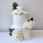 A two-tiered cake with a white frosting and decorated with silk roses, green leaves, and white accents.