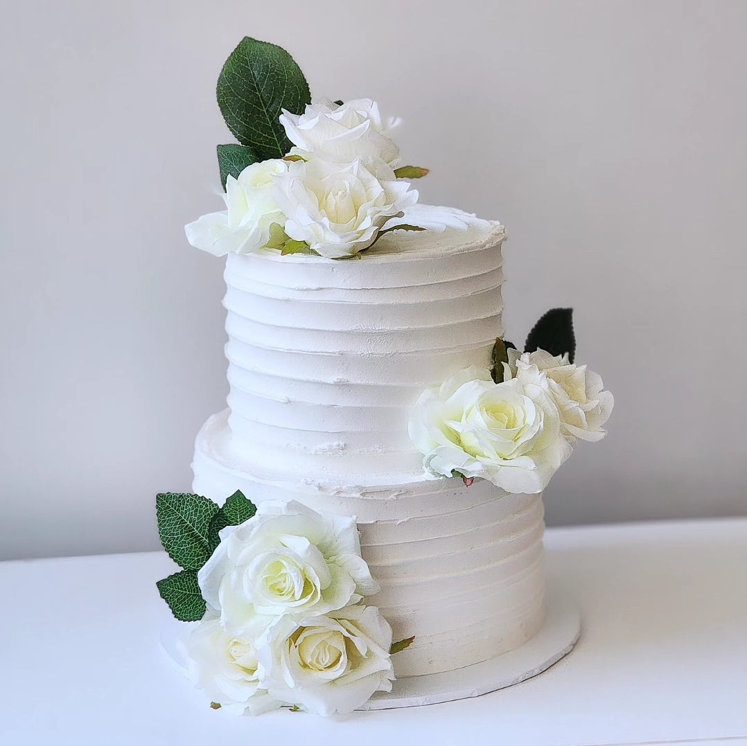 A two-tiered cake with a white frosting and decorated with silk roses, green leaves, and white accents.