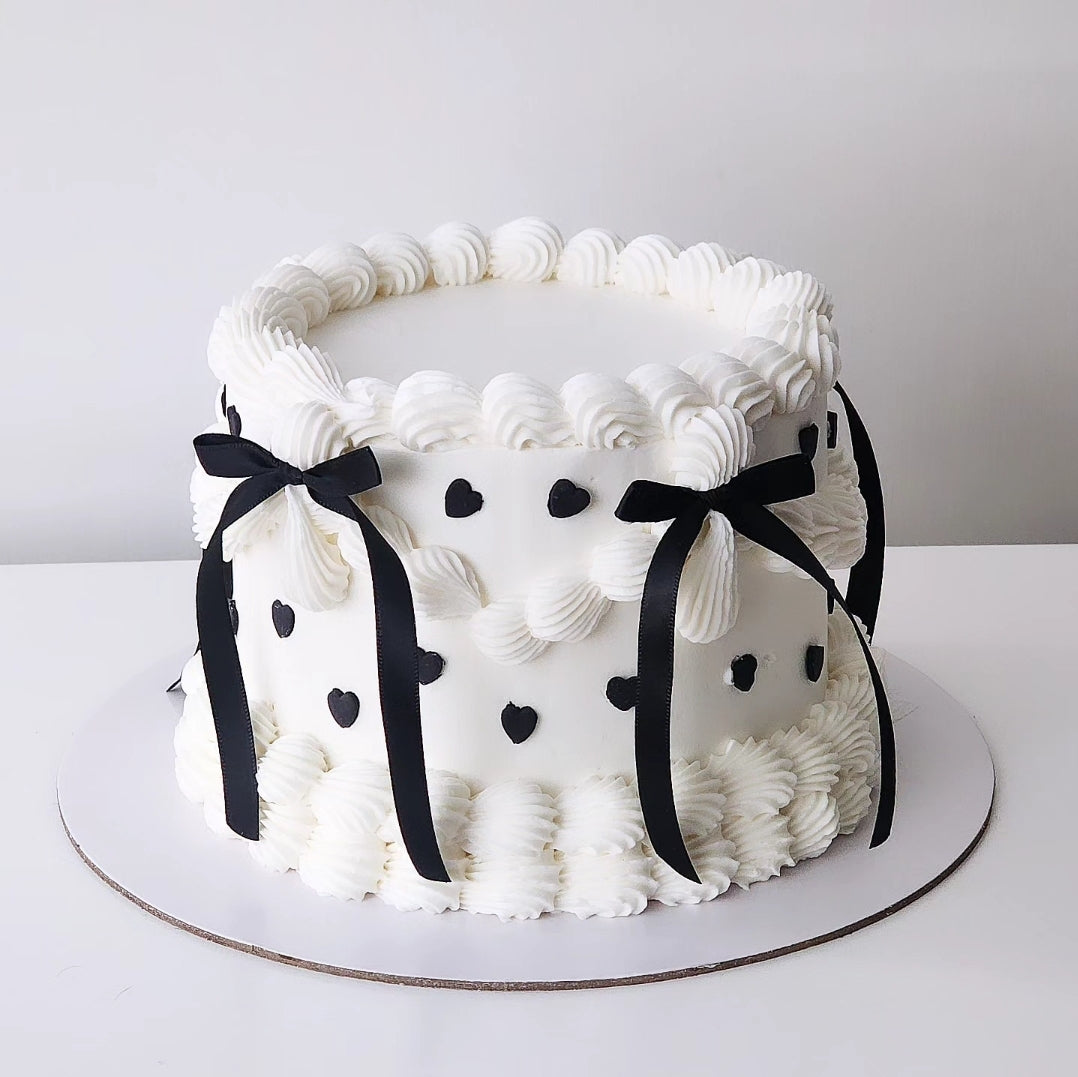 ALux with hearts Cake decorated with white frosting and black ribbons, with heart decorations on the sides.