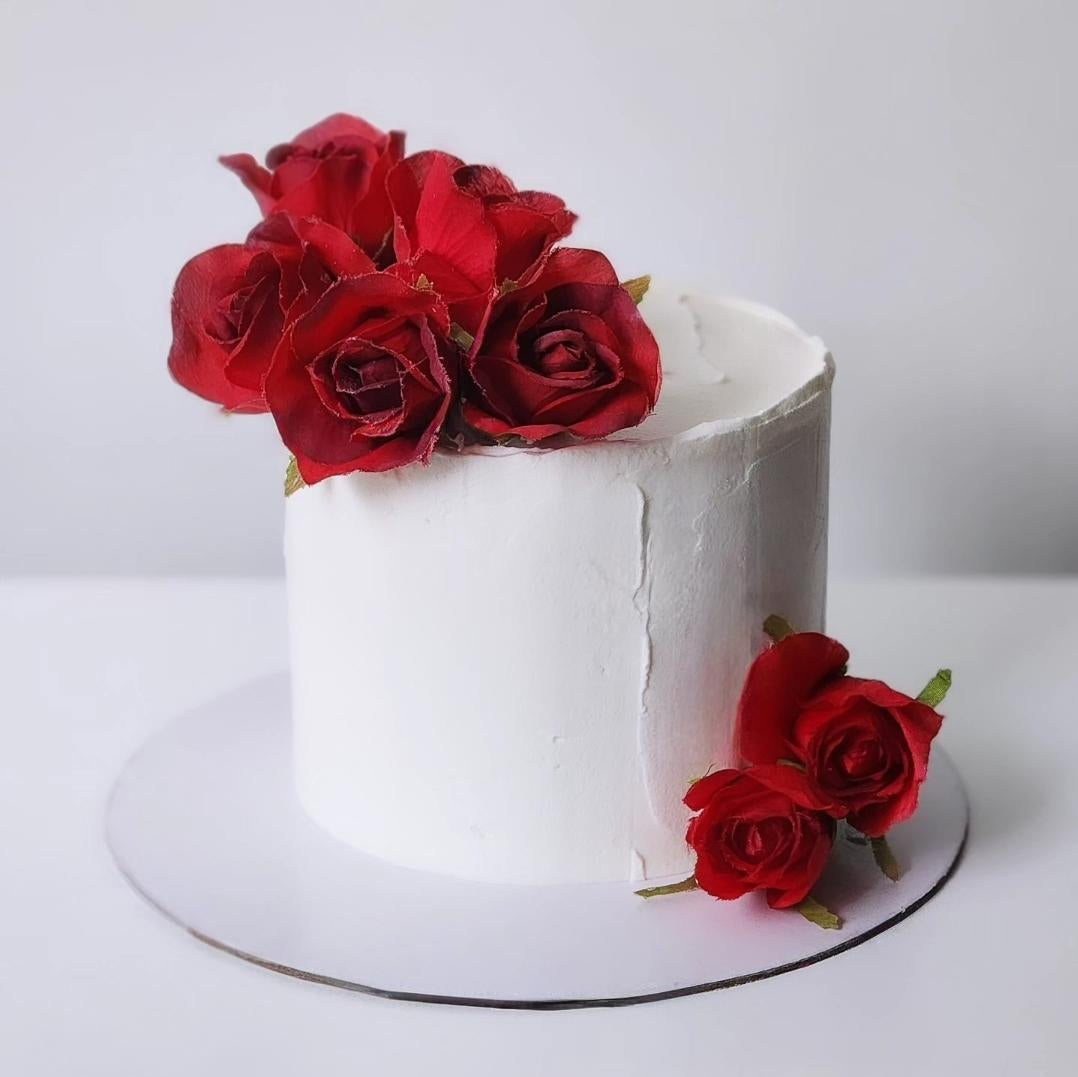 A decorative cake with a white sponge base and vanilla frosting, topped with red roses and green leaves, placed on a white surface.