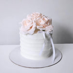 A layered cake with white frosting and pink artificial flowers, decorated with a white ribbon on top.