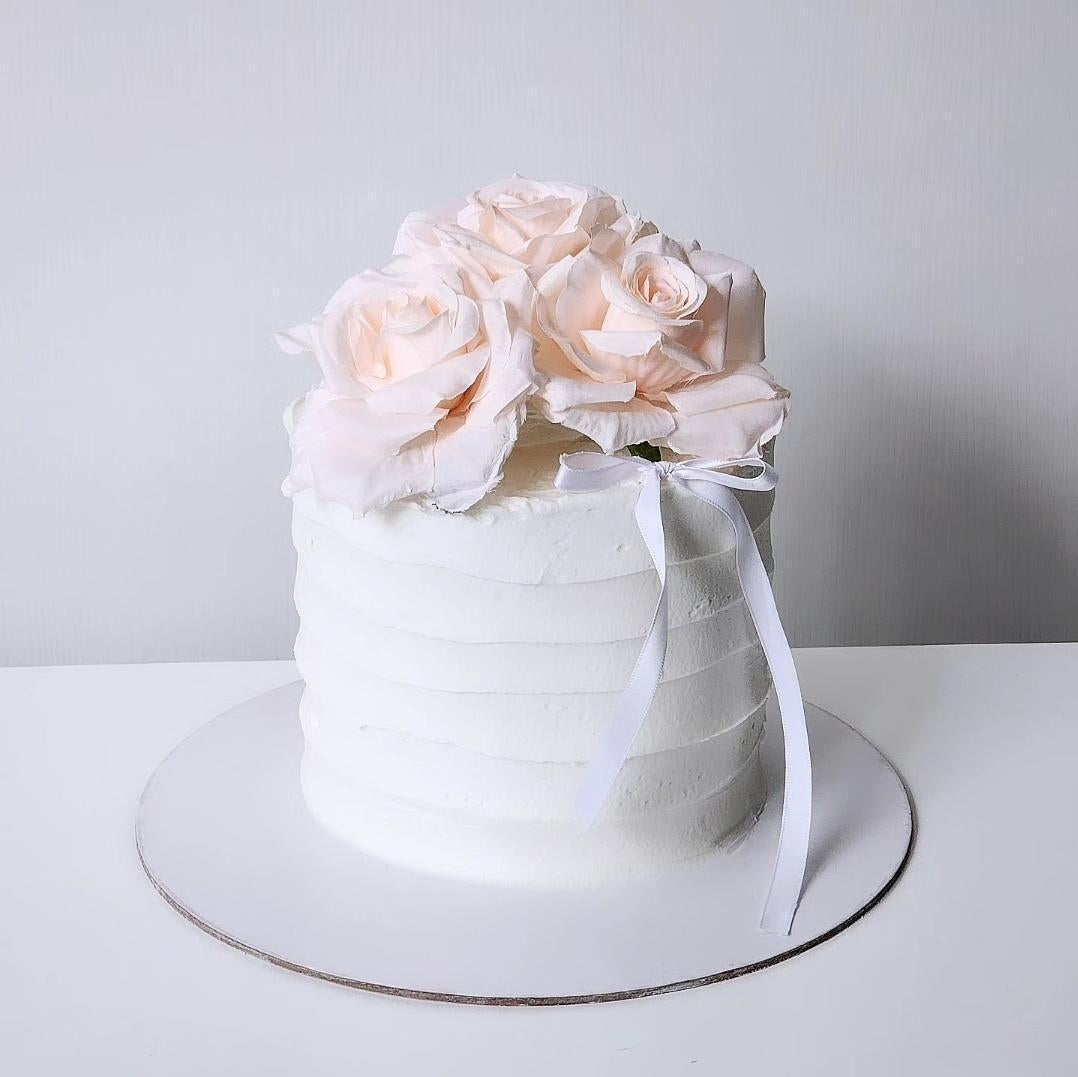 A layered cake with white frosting and pink artificial flowers, decorated with a white ribbon on top.