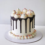 A celebration cake with white frosting and colorful sprinkles on top, with a chocolate glazed layer and a cream swirl on the side.