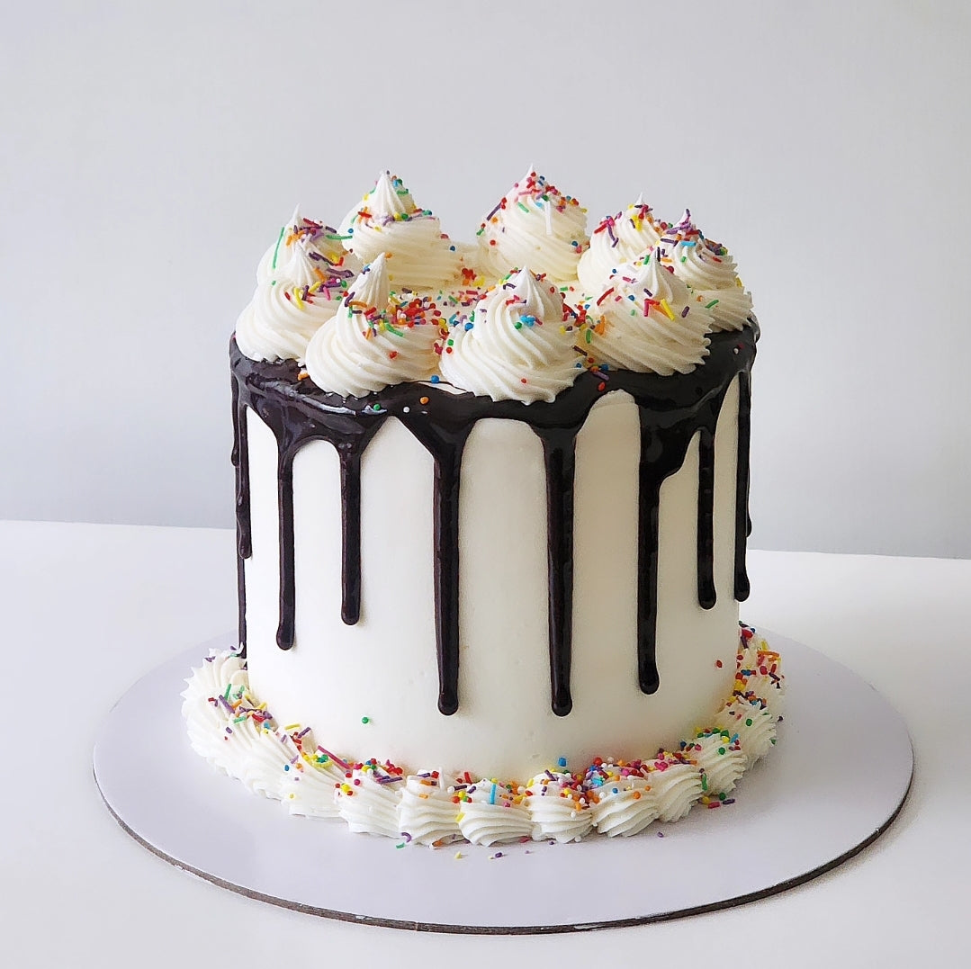 A celebration cake with white frosting and colorful sprinkles on top, with a chocolate glazed layer and a cream swirl on the side.