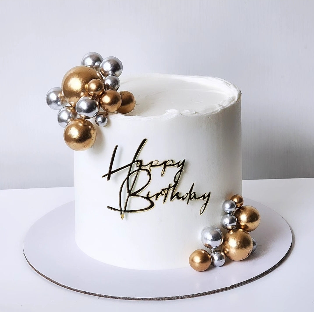 White birthday cake with gold and silver decorations and 'Happy Birthday' text on a white background