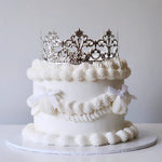 A cake with a white velvety frosting and an acrylic crown on top, decorated with satin ribbon.