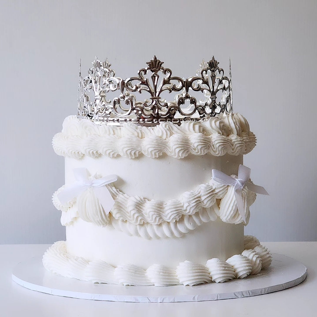 A cake with a white velvety frosting and an acrylic crown on top, decorated with satin ribbon.