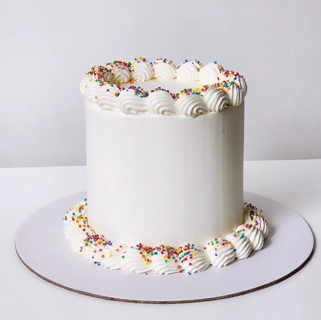 A decorated cake with white frosting and multicolored sprinkles on top, placed on a white cake stand.