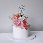 A decorated cake with white buttercream frosting and artificial pink and orange florals on top, presented on a white plate against a neutral background.