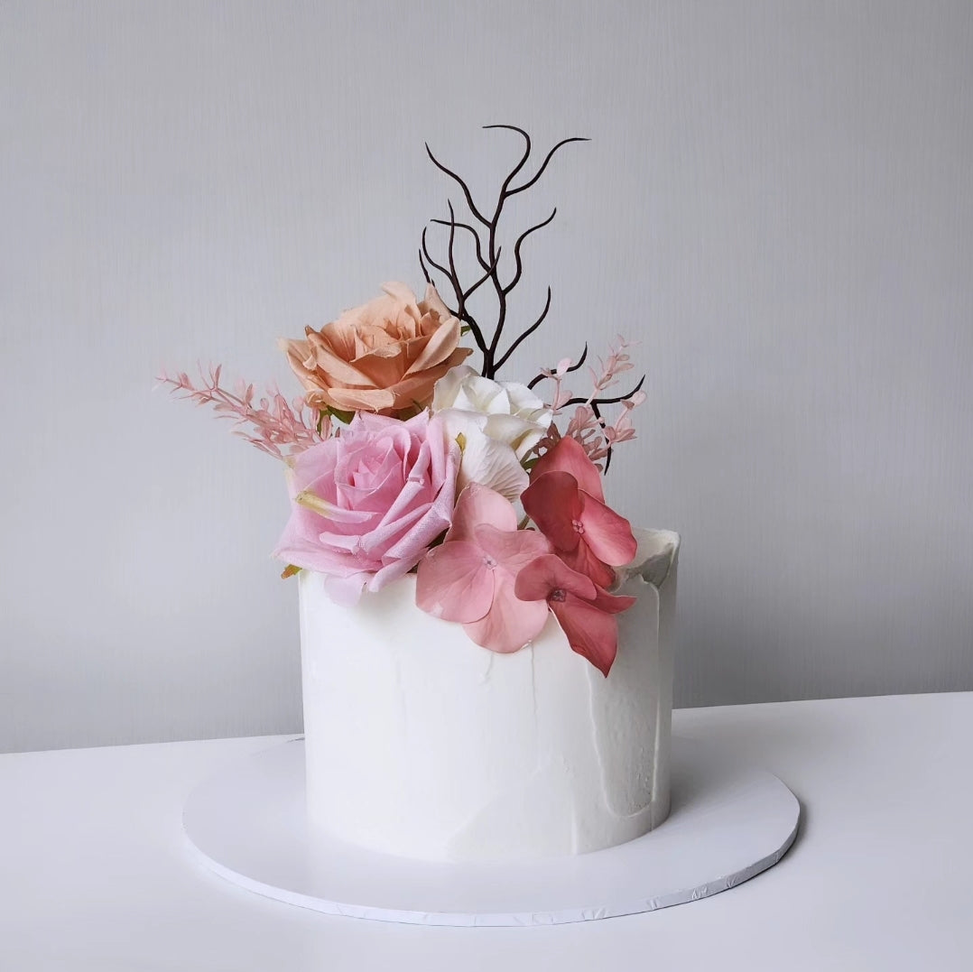 A decorated cake with white buttercream frosting and artificial pink and orange florals on top, presented on a white plate against a neutral background.