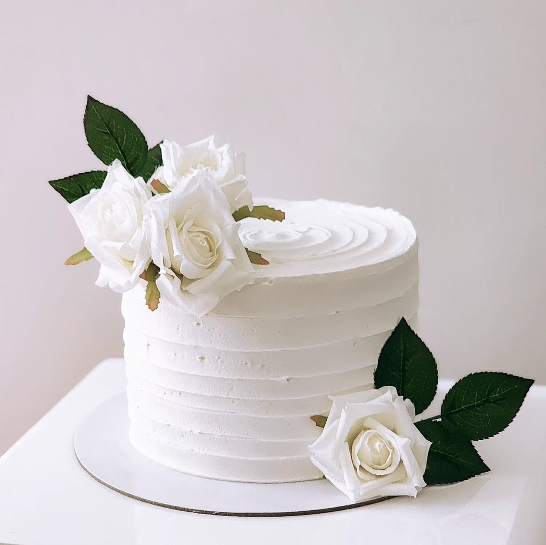 A white cake with a layer of white frosting and decorated with silk roses in a vase on top.