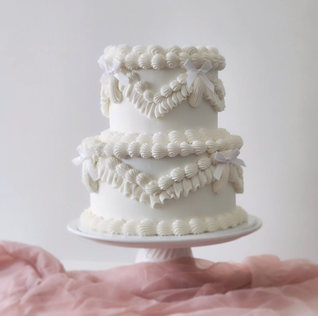 A two-tiered cake with a white frosting and decorative elements on a white cake stand against a pink-faded background.
