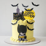 A themed birthday cake decorated to look like a Bat character with bats around, in yellow and black colors, on a white cake stand.