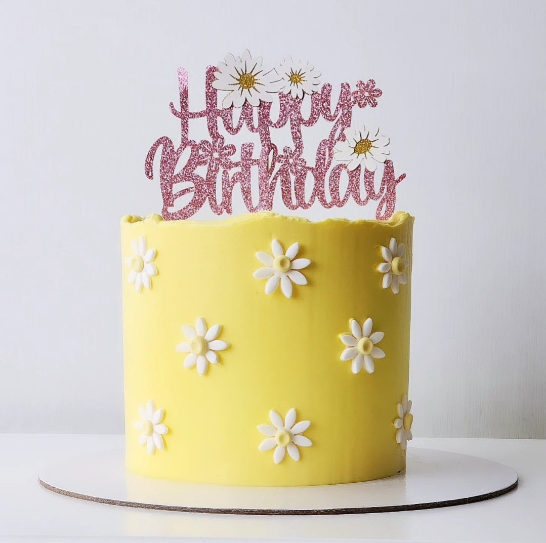 A yellow birthday cake with white daisy decorations and a pink 'Happy Birthday' cake topper.