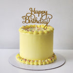 A yellow celebration cake with 'Happy Birthday' written on it, frosted with white buttercream and sprinkles on top.
