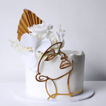 An elegant cake with a white sponge layer and golden decorative elements shaped as a stylized woman's face and hair, frosted with vanilla buttercream and adorned with white flowers.