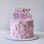 Pink birthday cake with white flowers and a 'Happy Birthday' topper on a white background