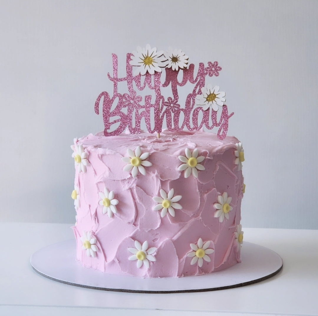 Pink birthday cake with white flowers and a 'Happy Birthday' topper on a white background