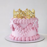 A decorated cake with pink buttercream frosting and a golden acrylic crown on top