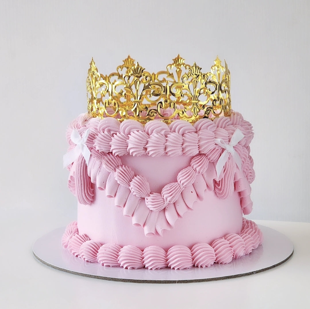 A decorated cake with pink buttercream frosting and a golden acrylic crown on top