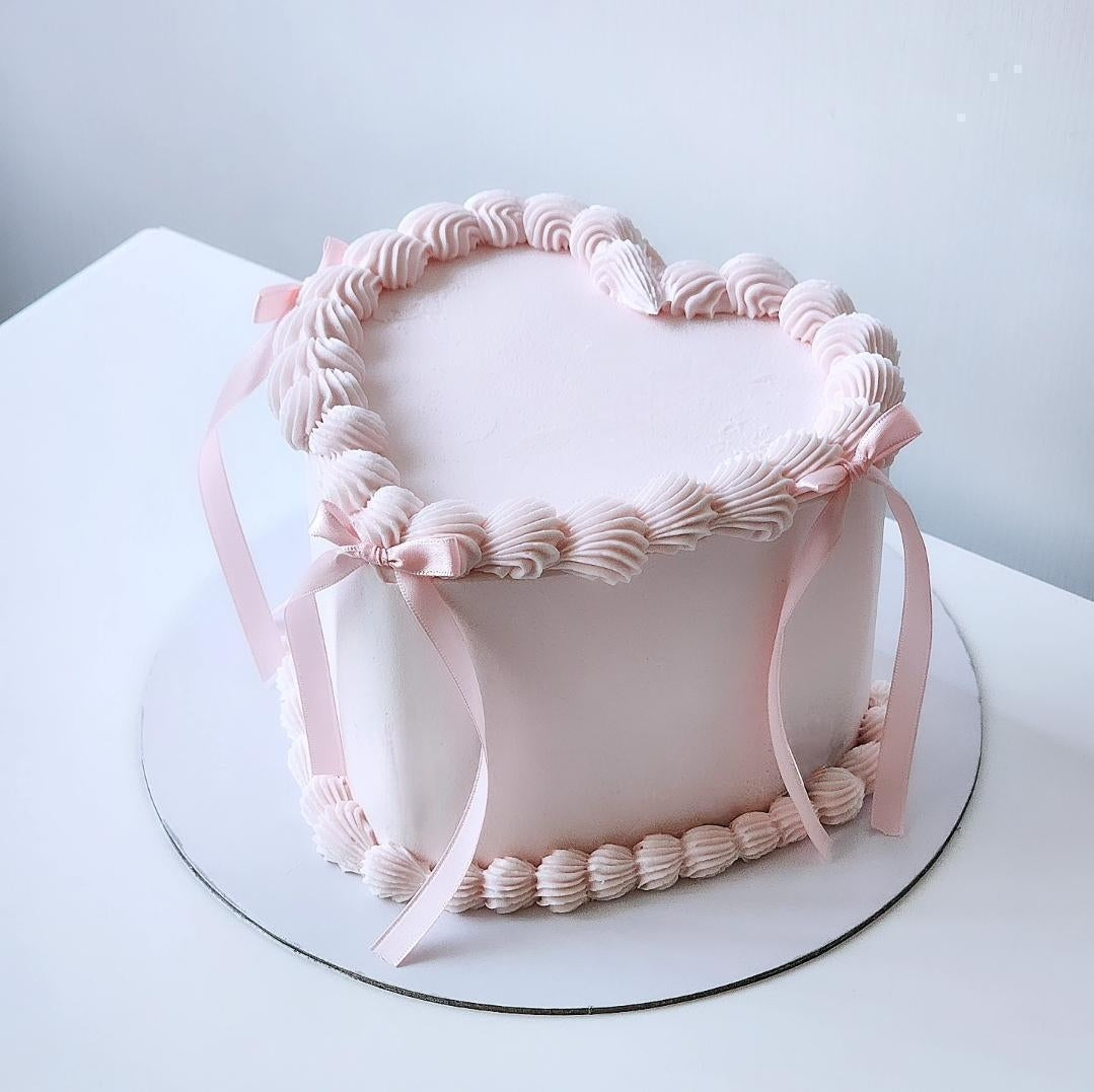 A heart-shaped cake decorated with pink buttercream and satin ribbons, on a white plate against a white background.