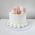 A layered cake with white frosting and four pink macarons on top, with a white cake stand and a plain background.