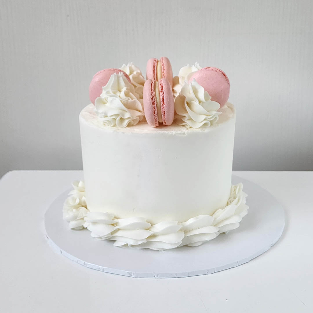 A layered cake with white frosting and four pink macarons on top, with a white cake stand and a plain background.
