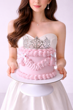 Woman holding a cake designed to look like a strapless dress with a silver brooch and pink ruffles.