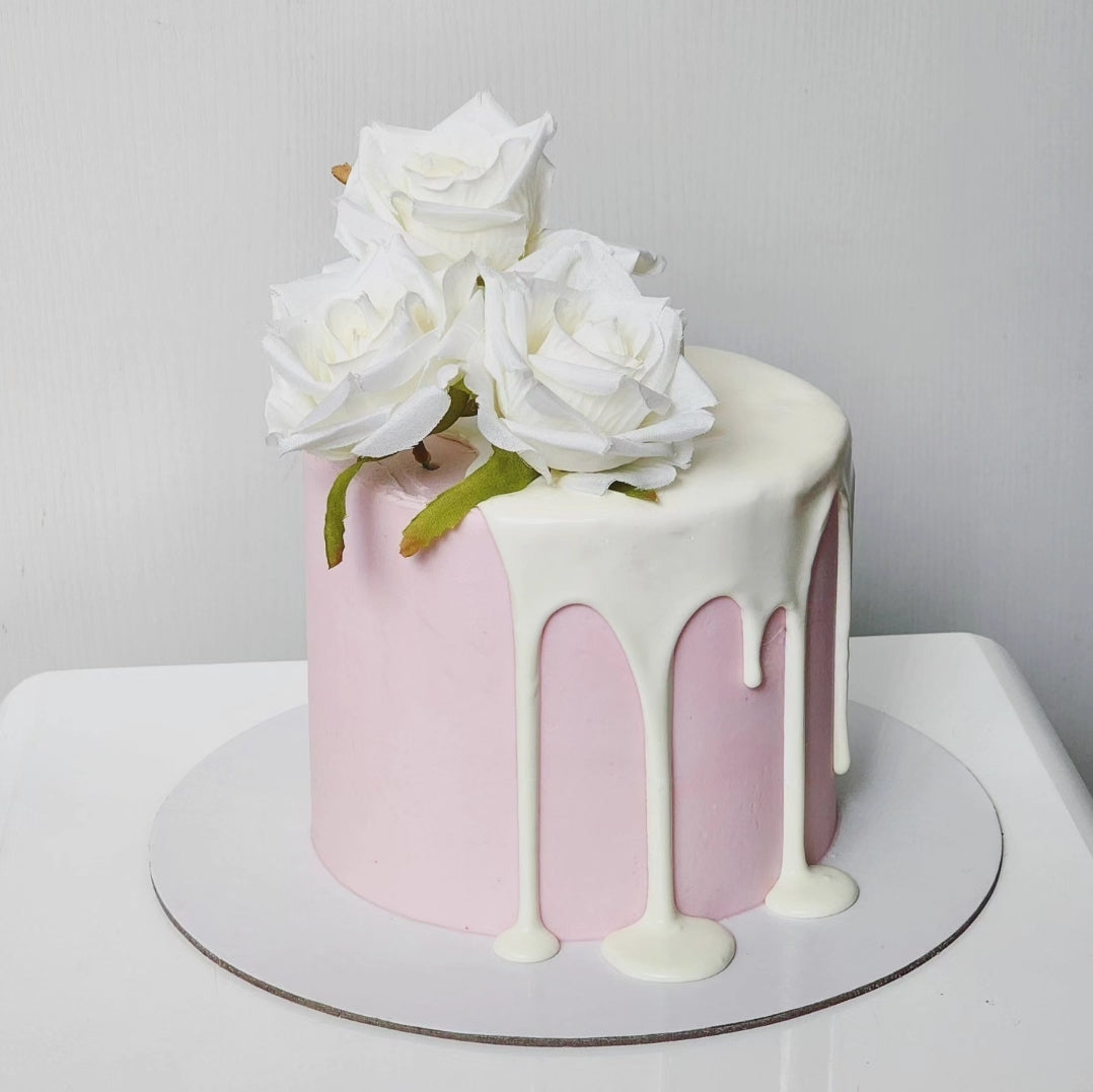 A pink cake with white frosting and a small white cake stand, topped with white silk roses and a creamy white chocolate ganache drizzle.
