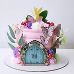 A decorative cake with a fairy-tale theme, featuring a pink base, green and purple decorations, a fairy door, and butterflies.