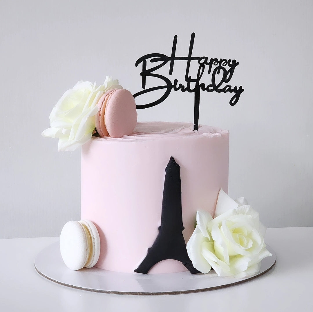 A birthday cake with a pink layer, a chocolate layer, and white frosting, decorated with a Eiffel Tower silhouette, a 'Happy Birthday' topper, white flowers, and a macaron.