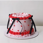 A layered cake with pink frosting, heart-shaped decorations, and a black ribbon on top, set against a white background.